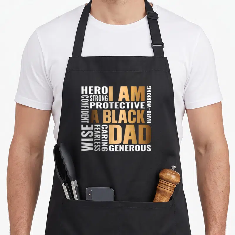 Matter father day apron - Gold and white typography reading I AM A BLACK DAD surrounded by adjectives HERO STRONG — shown on a black cotton canvas apron against a white background