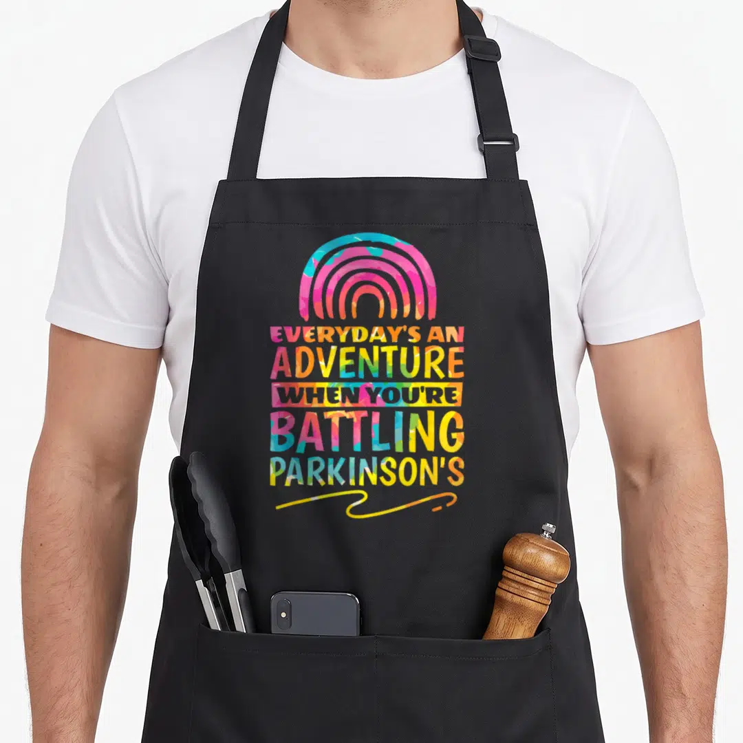 Warrior parkinsons awareness apron - Rainbow arch and stacked text reading Everyday's An Adventure When You're Battling — shown on a black cotton canvas apron against a white background