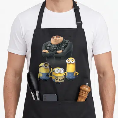 Despicable father day apron - Gru stands behind three Minions, one holding a #1 DAD cardboard sign — shown on a black cotton canvas apron against a white background
