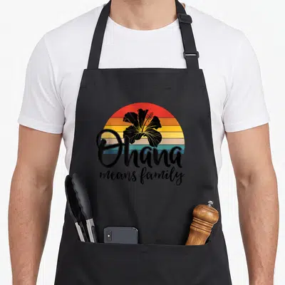 Ohana hawaiian apron - Black hibiscus on red orange yellow teal stripes with Ohana means family script text — shown on a black cotton canvas apron against a white background