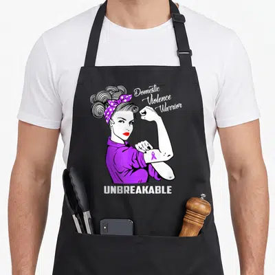 Violence domestic aprons - Cartoon woman flexing arm with purple headscarf and UNBREAKABLE text — shown on a black cotton canvas apron against a white background