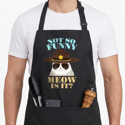 State cat apron - Cartoon cat in brown trooper hat with sunglasses and blue tan text — shown on a black cotton canvas apron against a white background