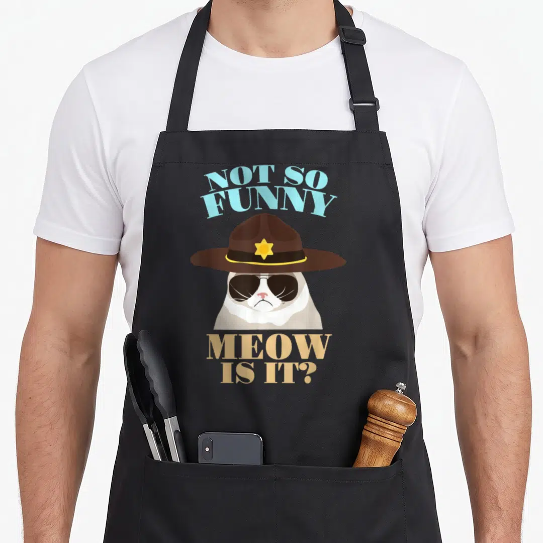 State cat apron - Cartoon cat in brown trooper hat with sunglasses and blue tan text — shown on a black cotton canvas apron against a white background