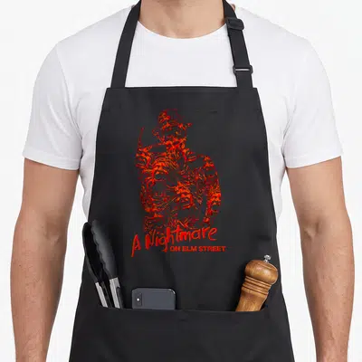 Chest elm apron - Distressed red Freddy Krueger illustration with A Nightmare ON ELM STREET text below — shown on a black cotton canvas apron against a white background