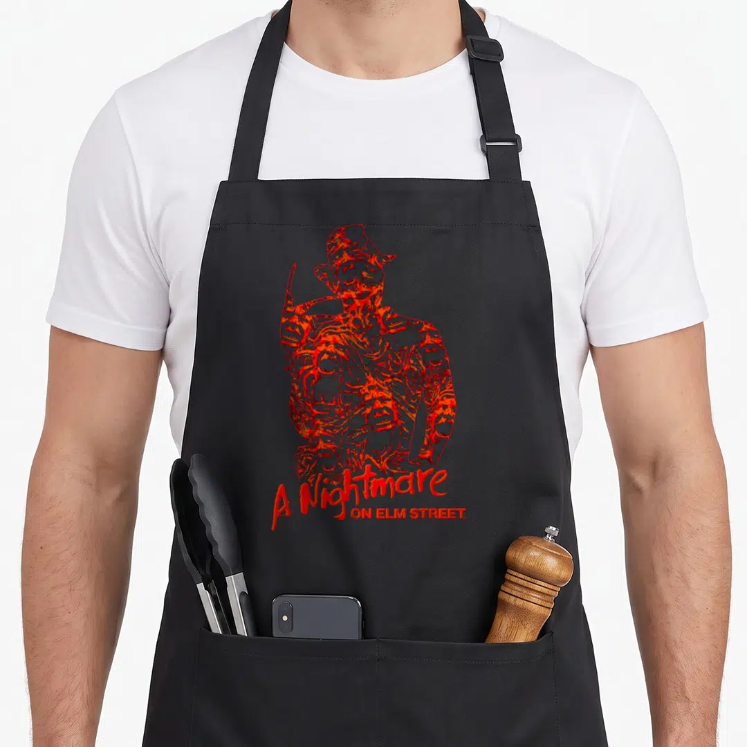 Chest elm apron - Distressed red Freddy Krueger illustration with A Nightmare ON ELM STREET text below — shown on a black cotton canvas apron against a white background