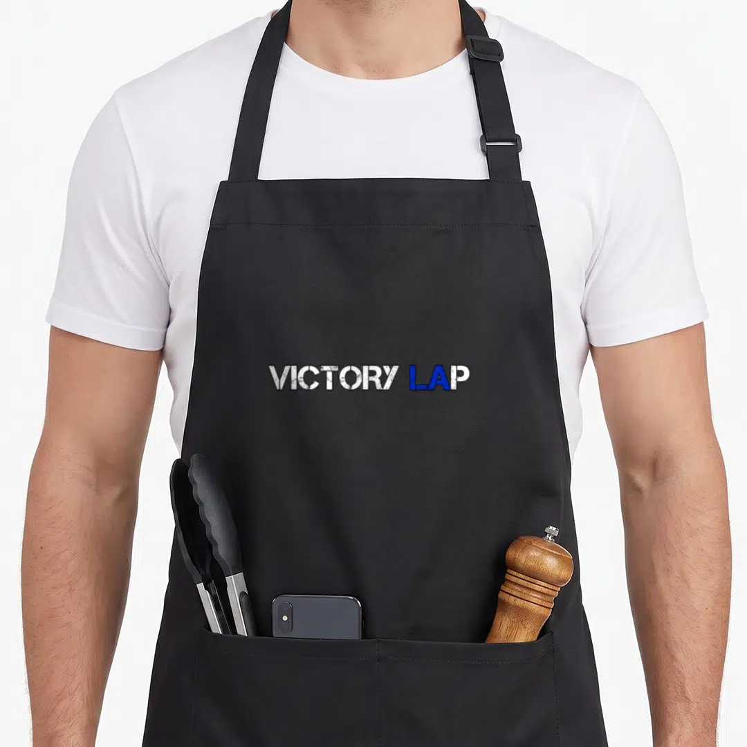 Cute trend apron - VICTORY LAP text in distressed white and blue block letters — shown on a black cotton canvas apron against a white background