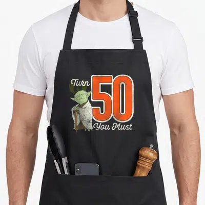 Yoda birthday apron - Cartoon Yoda stands next to large orange 50 with white text reading Turn You Must — shown on a black cotton canvas apron against a white background