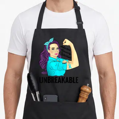 Unbreakable addiction recovery apron - Cartoon woman with purple hair flexing arm showing blue ribbon and Addiction — shown on a black cotton canvas apron against a white background