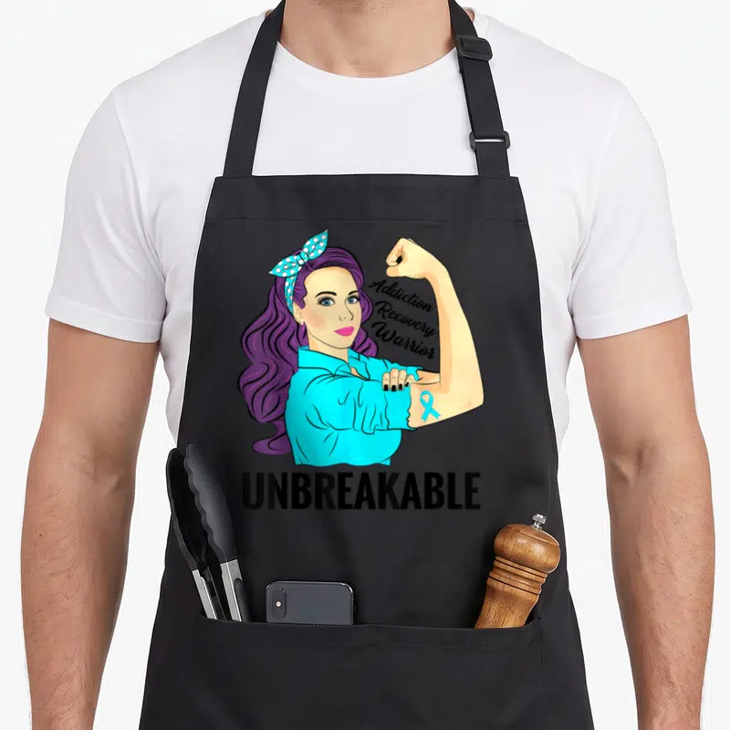 Unbreakable addiction recovery apron - Cartoon woman with purple hair flexing arm showing blue ribbon and Addiction — shown on a black cotton canvas apron against a white background