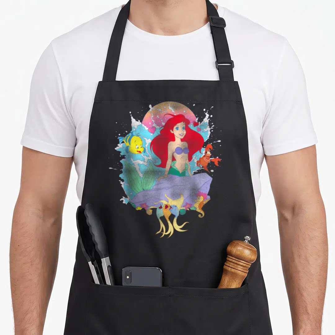 Ariel splash aprons - Cartoon mermaid on grey rock with yellow fish, orange crab, pink hearts, and colorful sun splashes — shown on a black cotton canvas apron against a white background