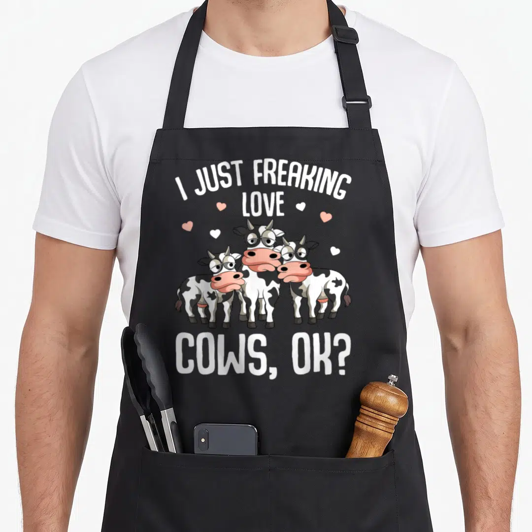 Freaking farmer apron - Three cartoon cows with pink noses and scattered hearts under the text I Just Freaking Love — shown on a black cotton canvas apron against a white background