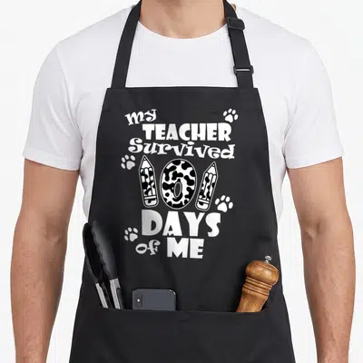 Survived teacher apron - Stacked text my TEACHER Survived 100 DAYS of ME with spotted number 100, pencils and paw — shown on a black cotton canvas apron against a white background