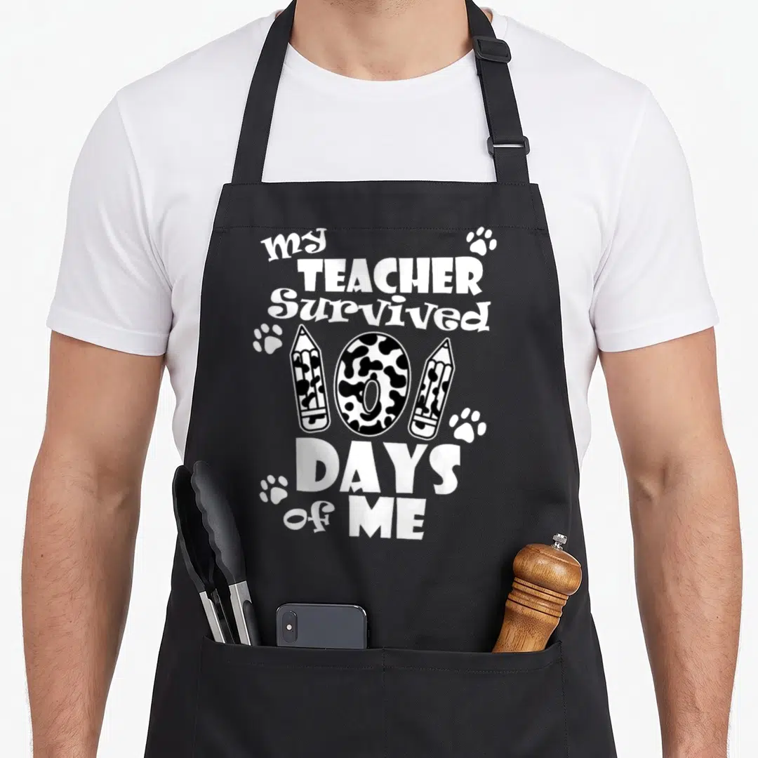 Survived teacher apron - Stacked text my TEACHER Survived 100 DAYS of ME with spotted number 100, pencils and paw — shown on a black cotton canvas apron against a white background