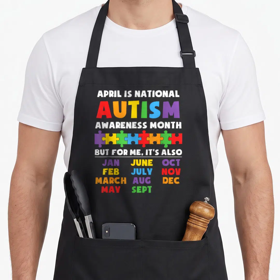 Support autism dad apron - Rainbow puzzle pieces separating white awareness text from a list of twelve colored month — shown on a black cotton canvas apron against a white background