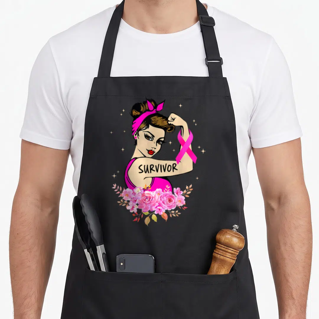 Rosie breast cancer awareness aprons - Cartoon woman flexing arm with SURVIVOR text, pink ribbon, and flowers — shown on a black cotton canvas apron against a white background