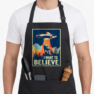 Believe area bigfoot apron - Retro poster art of a UFO abducting a Bigfoot silhouette with text I WANT TO BELIEVE below — shown on a black cotton canvas apron against a white background
