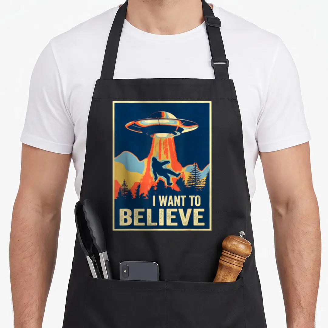 Believe area bigfoot apron - Retro poster art of a UFO abducting a Bigfoot silhouette with text I WANT TO BELIEVE below — shown on a black cotton canvas apron against a white background