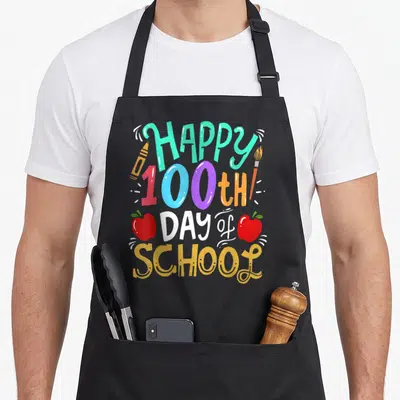 Cute 100th teacher apron - Colorful HAPPY 100th DAY of SCHOOL text with crayon, paintbrush, and two red apples — shown on a black cotton canvas apron against a white background
