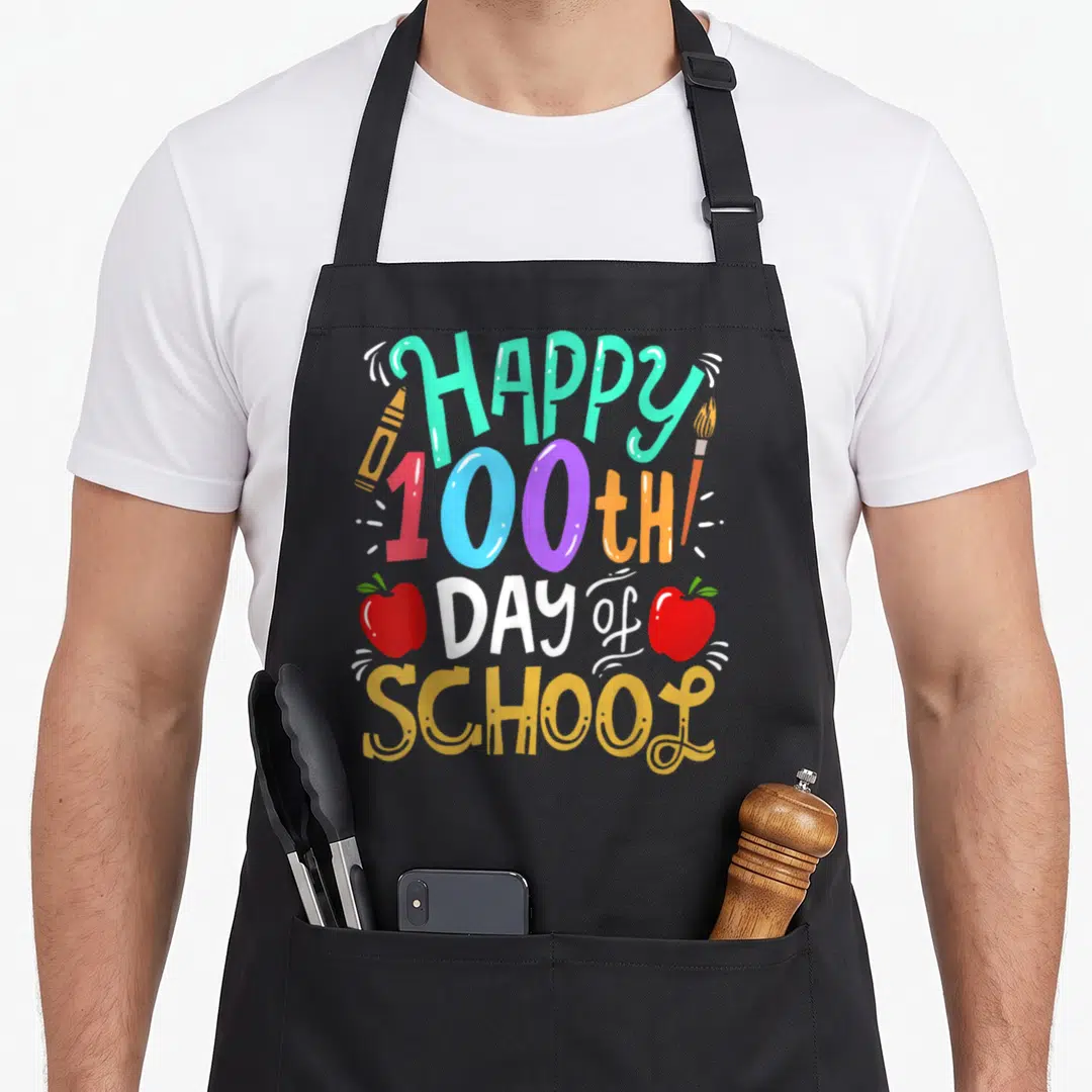 Cute 100th teacher apron - Colorful HAPPY 100th DAY of SCHOOL text with crayon, paintbrush, and two red apples — shown on a black cotton canvas apron against a white background