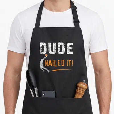 Basketball dude apron - DUDE NAILED IT text with jumping basketball player silhouette and orange swoosh — shown on a black cotton canvas apron against a white background