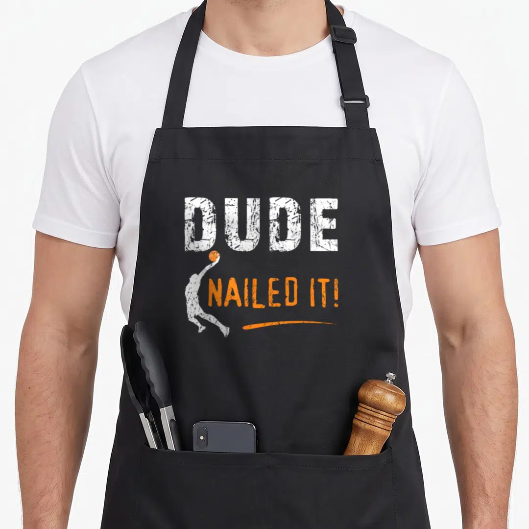 Basketball dude apron - DUDE NAILED IT text with jumping basketball player silhouette and orange swoosh — shown on a black cotton canvas apron against a white background