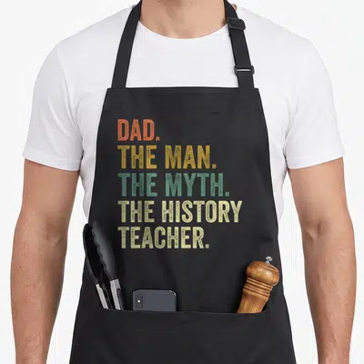 History father day apron - Stacked text DAD THE MAN THE MYTH THE HISTORY TEACHER in orange yellow green beige — shown on a black cotton canvas apron against a white background