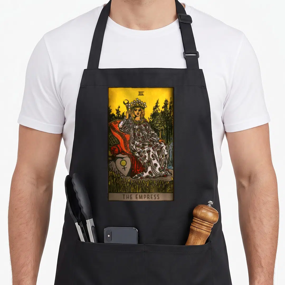 Empress tarot card apron - Realistic woman on throne with scepter, heart symbol, and THE EMPRESS text in yellow, red — shown on a black cotton canvas apron against a white background
