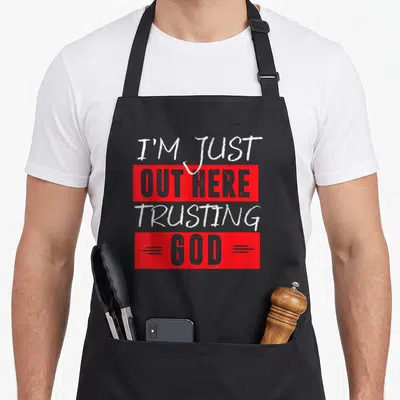 Trusting novelty apron - Stacked text I'm Just Out Here Trusting God in red white grey blocks — shown on a black cotton canvas apron against a white background
