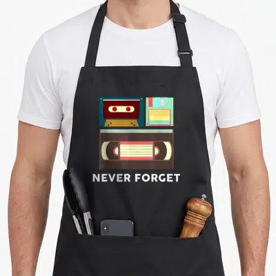 Forget housewarming apron - Cassette tape, floppy disk, and VHS tape illustration with Never Forget text in blue — shown on a black cotton canvas apron against a white background