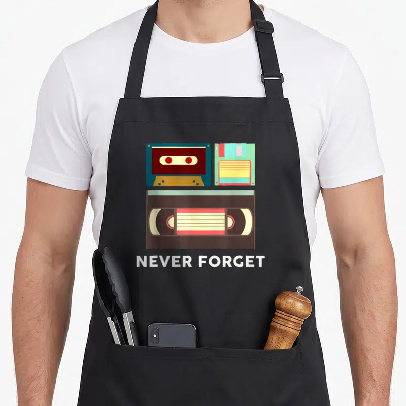 Forget housewarming apron - Cassette tape, floppy disk, and VHS tape illustration with Never Forget text in blue — shown on a black cotton canvas apron against a white background
