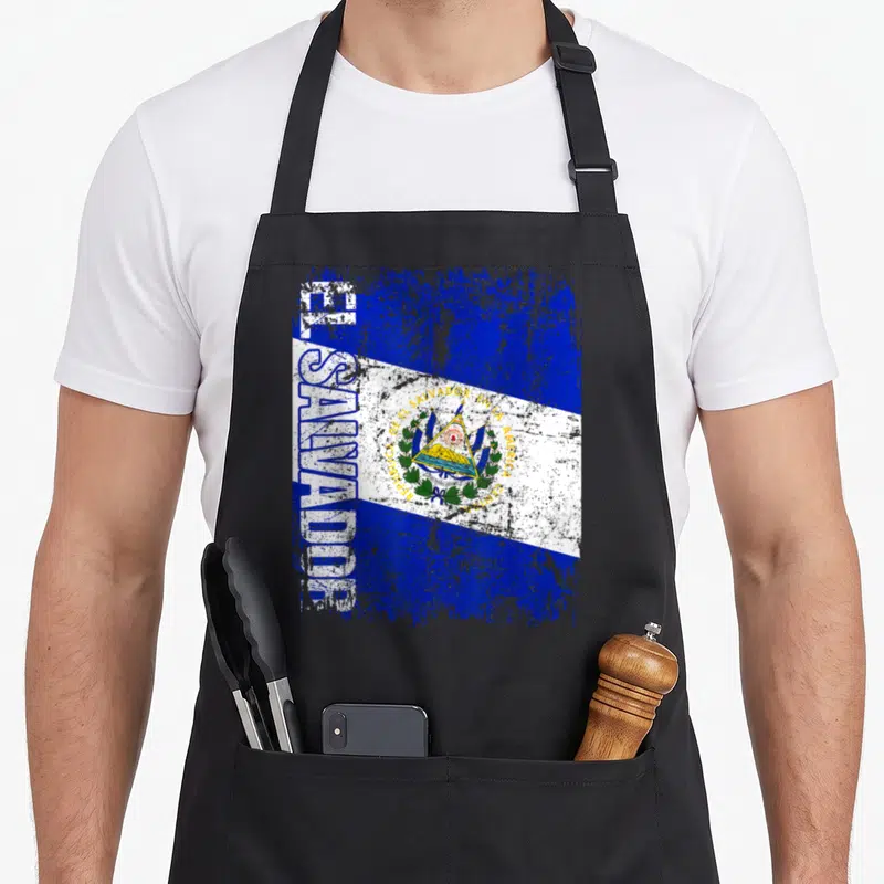 Distressed el cook apron - Vertical EL SALVADOR text beside blue white distressed flag coat of arms — shown on a black cotton canvas apron against a white background