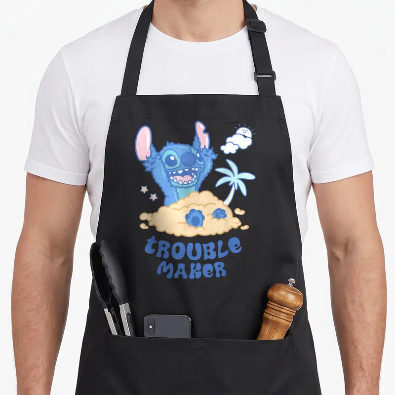 Stitch maker apron - Blue cartoon character peeking from tan sand with Troublemaker text and palm tree — shown on a black cotton canvas apron against a white background