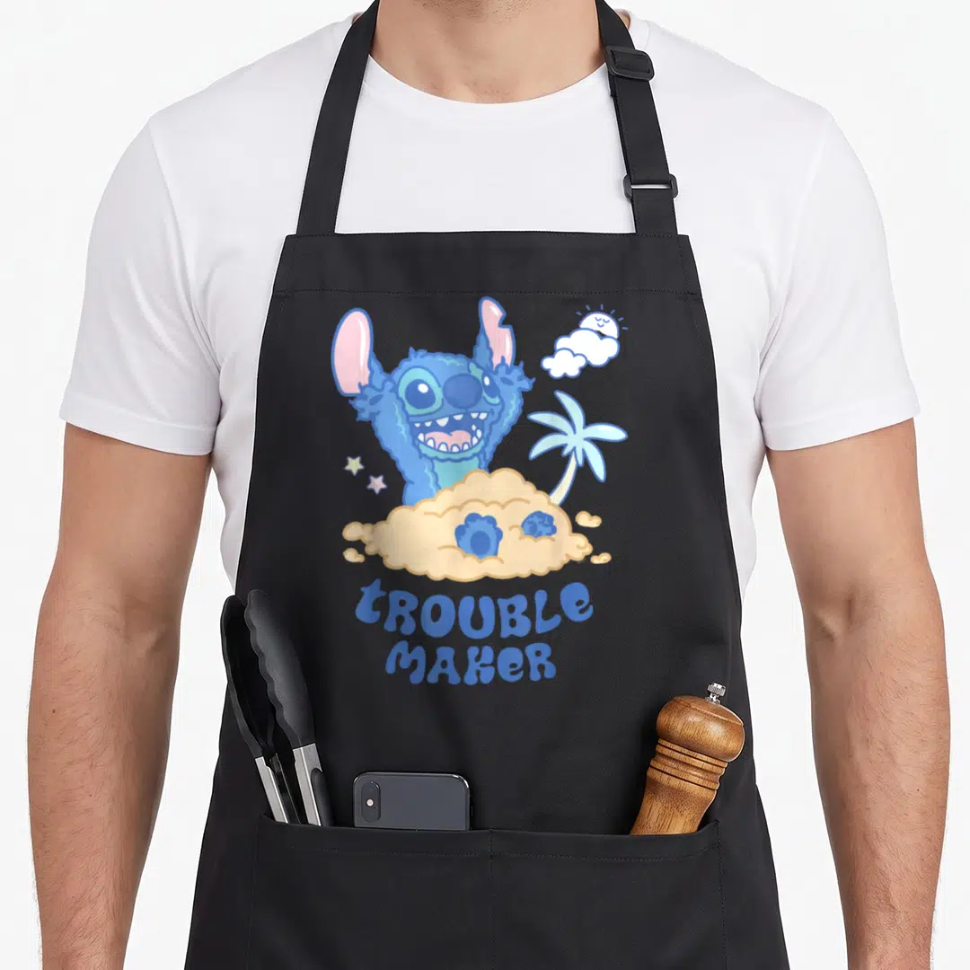 Stitch maker apron - Blue cartoon character peeking from tan sand with Troublemaker text and palm tree — shown on a black cotton canvas apron against a white background