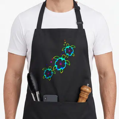 Honu tropical print aprons - Three cartoon sea turtles with flowers on shells, diagonal layout, blue green yellow pink — shown on a black cotton canvas apron against a white background
