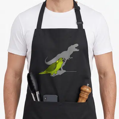 Birb dinosaur apron - Green cartoon parrot standing before a faint grey dinosaur silhouette — shown on a black cotton canvas apron against a white background