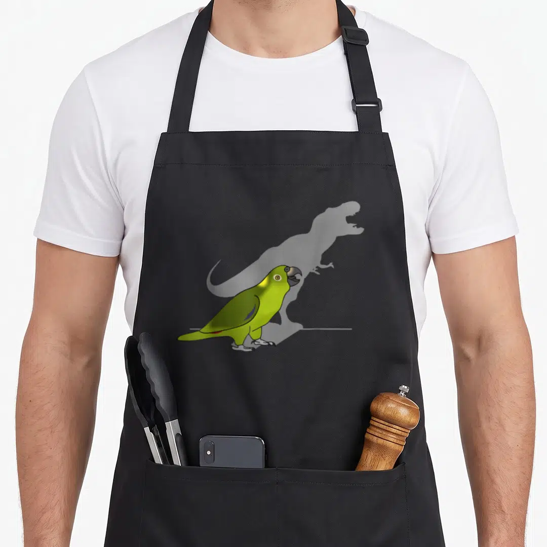 Birb dinosaur apron - Green cartoon parrot standing before a faint grey dinosaur silhouette — shown on a black cotton canvas apron against a white background