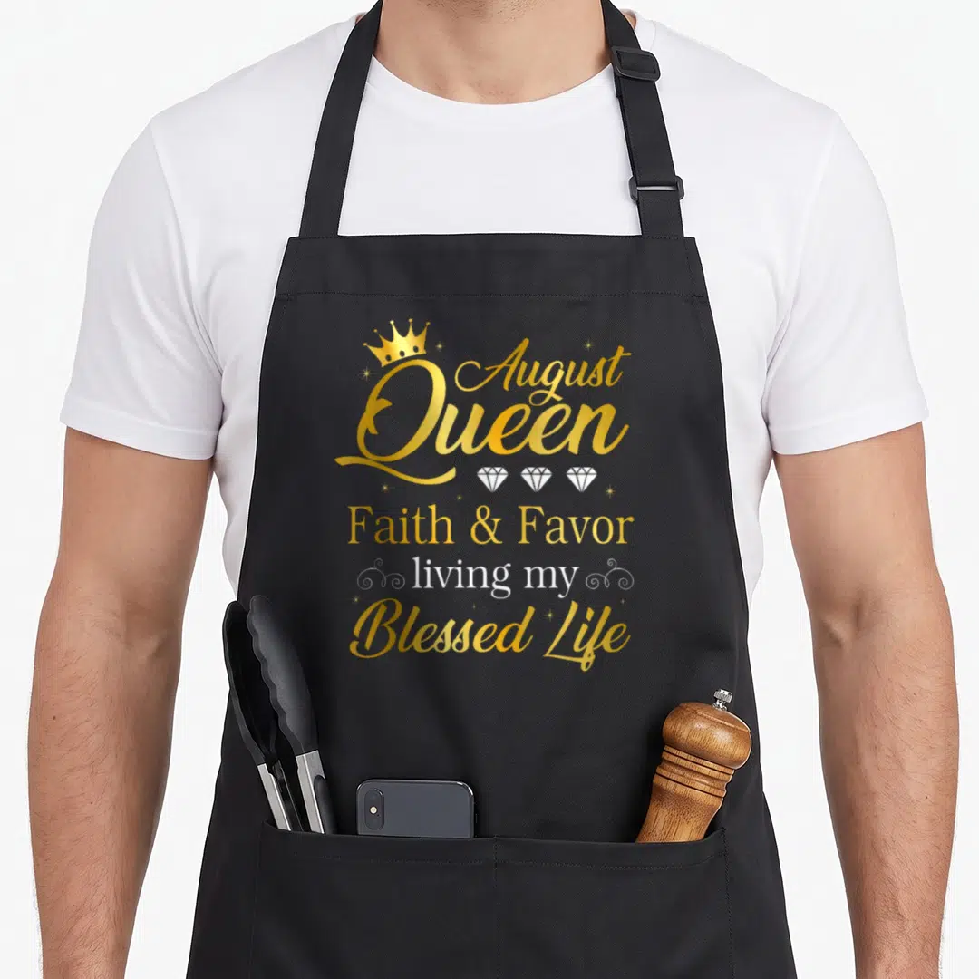 Living birthday apron - Gold and white text reading August Queen Faith Favor living my Blessed Life with crown — shown on a black cotton canvas apron against a white background