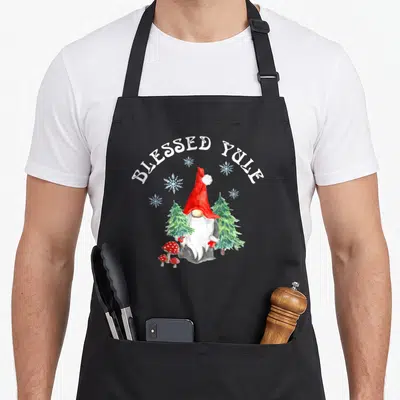 Solstice holiday apron - Watercolor gnome with red hat, pine trees, mushrooms, snowflakes, and arched BLESSED YULE text — shown on a black cotton canvas apron against a white background