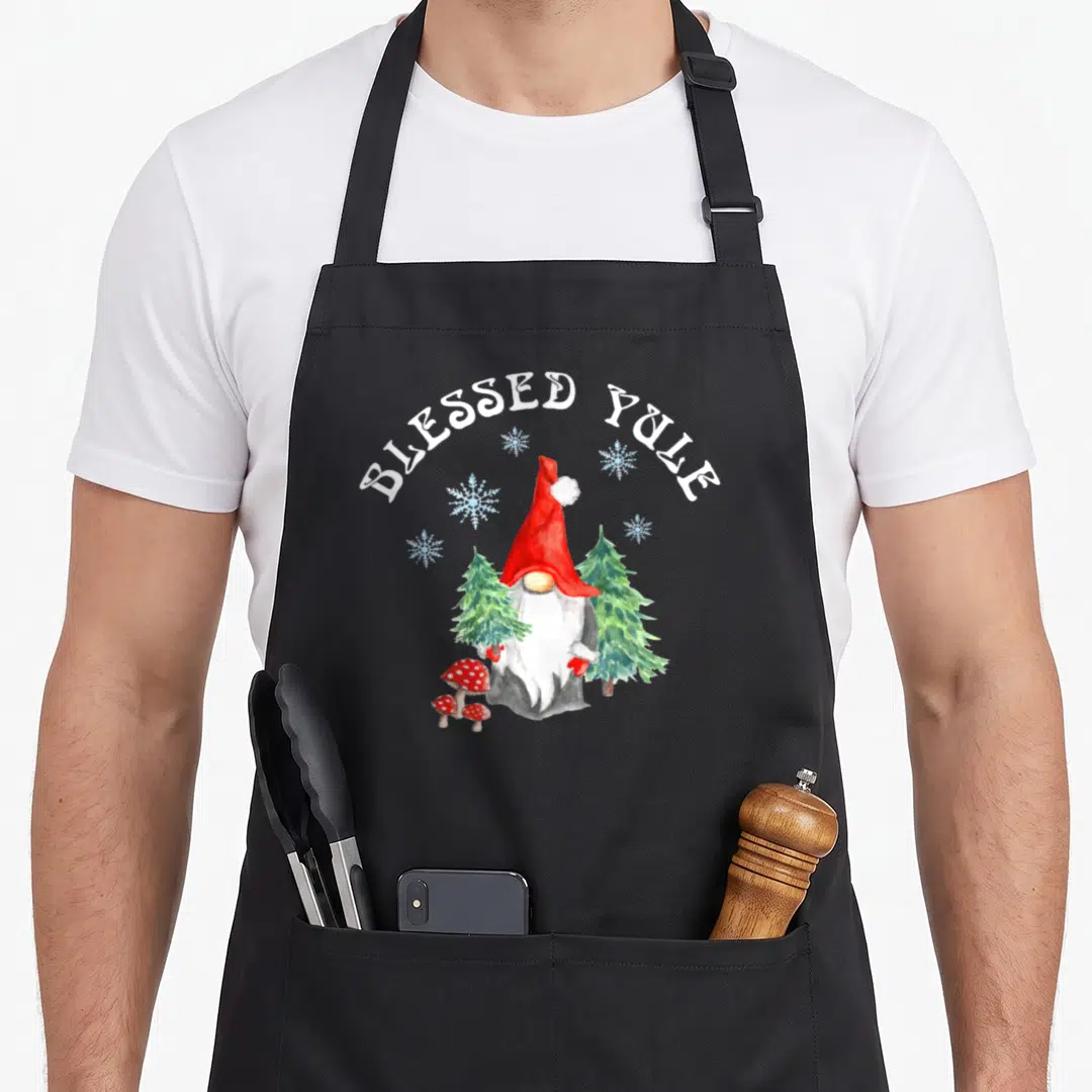 Solstice holiday apron - Watercolor gnome with red hat, pine trees, mushrooms, snowflakes, and arched BLESSED YULE text — shown on a black cotton canvas apron against a white background