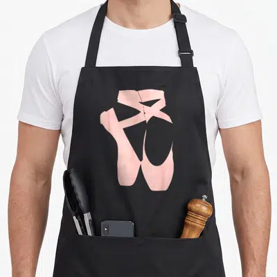 Dancing ballerina apron - Two pink ballet shoes with crossing ribbons on black bib — shown on a black cotton canvas apron against a white background