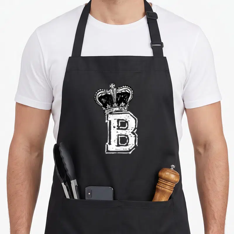 Queen bee apron - Black crown sitting on top of a large white distressed letter B in line art — shown on a black cotton canvas apron against a white background