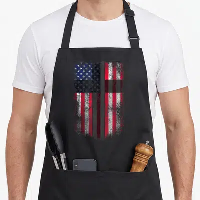 Flag cross christian aprons - Distressed American flag design with a vertical cross in red white blue and grey — shown on a black cotton canvas apron against a white background