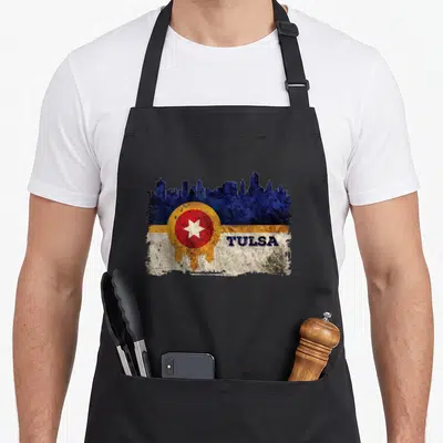 Skyline distressed apron - Blue Tulsa skyline above yellow band, red circle white star, distressed text TULSA — shown on a black cotton canvas apron against a white background