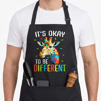 Acceptance giraffe apron - Mosaic giraffe head with puzzle pieces and IT'S OKAY TO BE DIFFERENT text — shown on a black cotton canvas apron against a white background