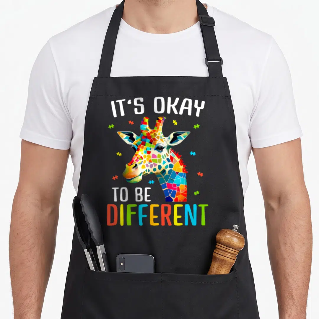 Acceptance giraffe apron - Mosaic giraffe head with puzzle pieces and IT'S OKAY TO BE DIFFERENT text — shown on a black cotton canvas apron against a white background