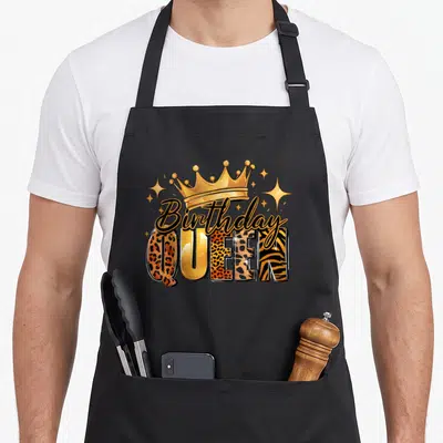 Afro queen of baking apron - Gold crown, script Birthday text, patterned QUEEN letters, and scattered stars — shown on a black cotton canvas apron against a white background