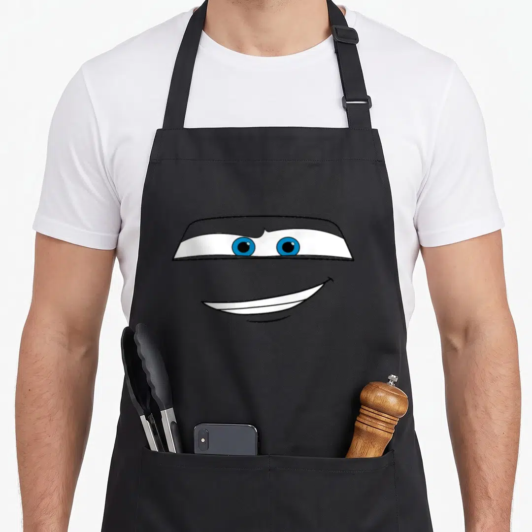 Face car apron - Cartoon grey face with blue eyes over a white mask and smiling mouth — shown on a black cotton canvas apron against a white background