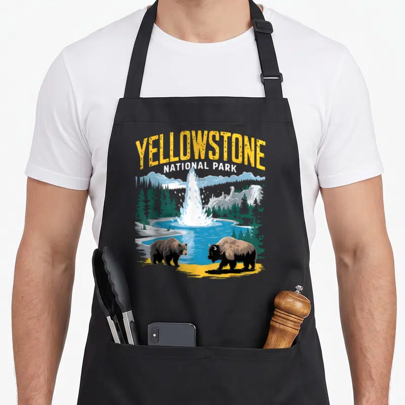 Bison bear apron - Retro Yellowstone National Park print with geyser, bear, and bison in yellow blue green and brown — shown on a black cotton canvas apron against a white background