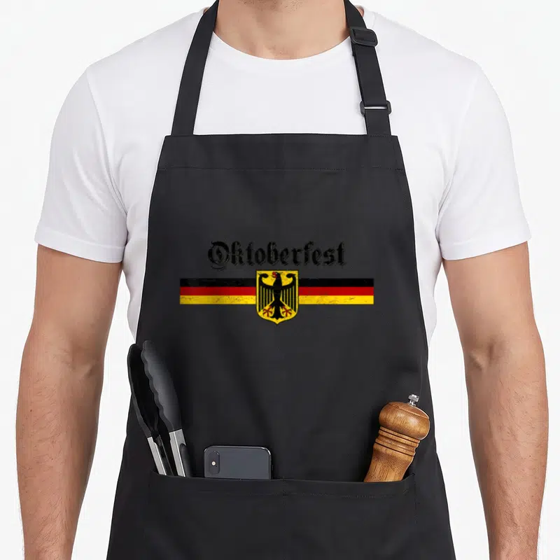 Coat german apron - Distressed Oktoberfest text above black red yellow flag and black eagle shield — shown on a black cotton canvas apron against a white background