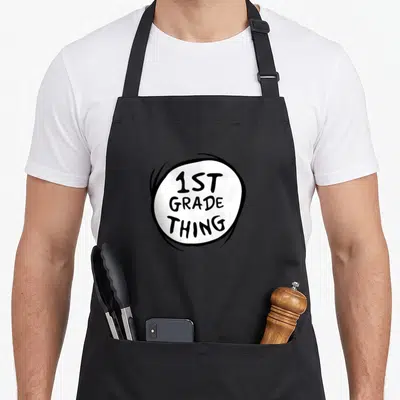 1st grade apron - White circle with black hand-drawn border containing stacked 1ST GRADE THING text — shown on a black cotton canvas apron against a white background